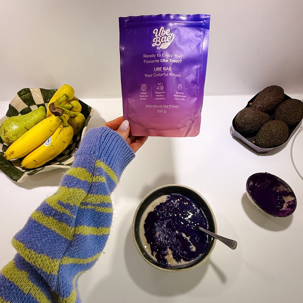 Hand holding Ube Bae organic ube powder pouch above a vibrant purple ube smoothie bowl with fresh bananas and avocados — real lifestyle UGC photo
