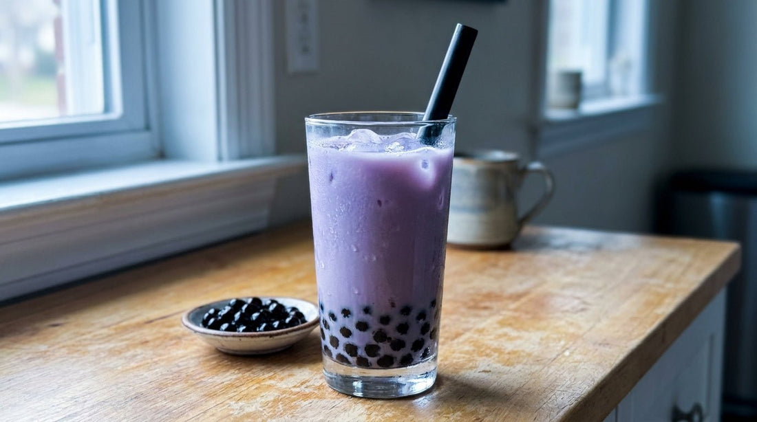 A tall glass of ube boba — creamy lavender-purple milk drink with black tapioca pearls settled at the bottom and a wide black boba straw inserted, condensation on the glass