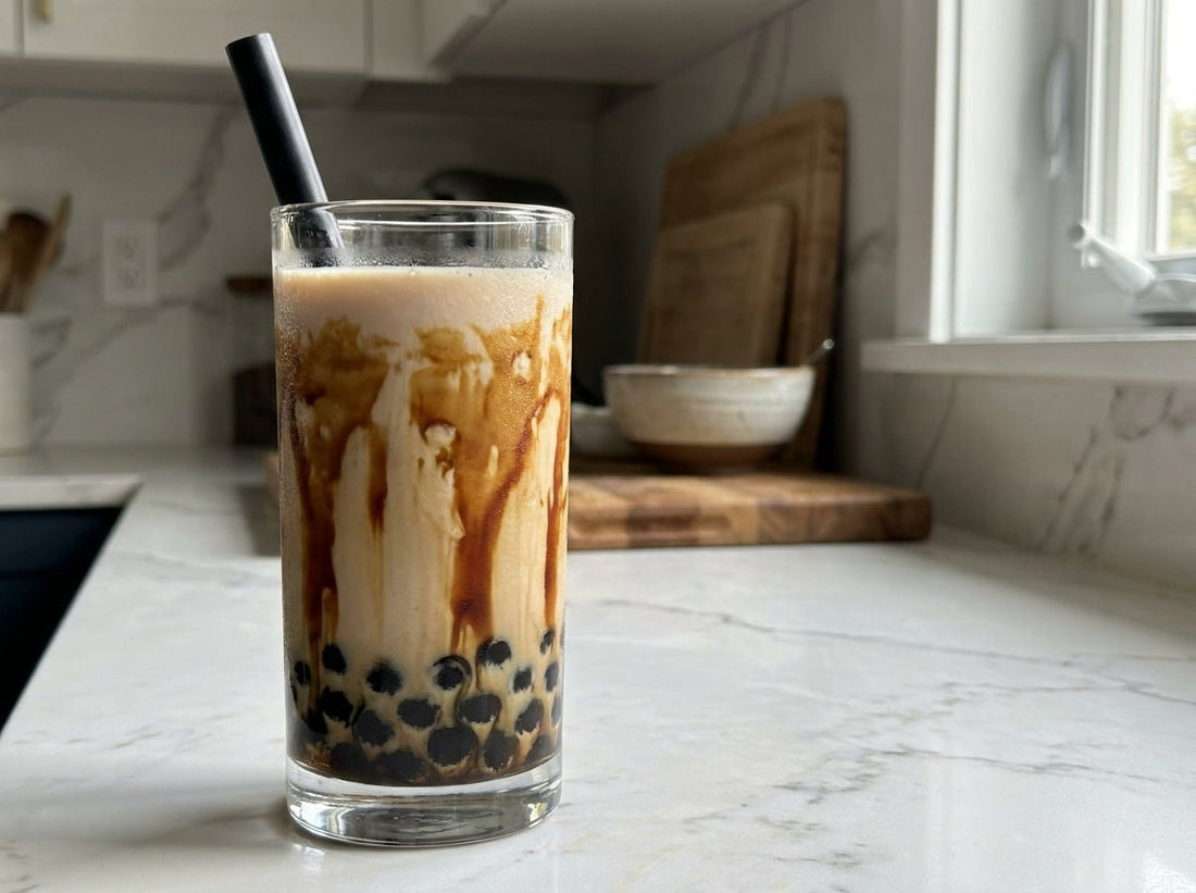 Tall clear glass of homemade ube boba with creamy lavender milk, dark brown sugar tiger stripes along the glass, black tapioca pearls clustered at the bottom, wide black boba straw
