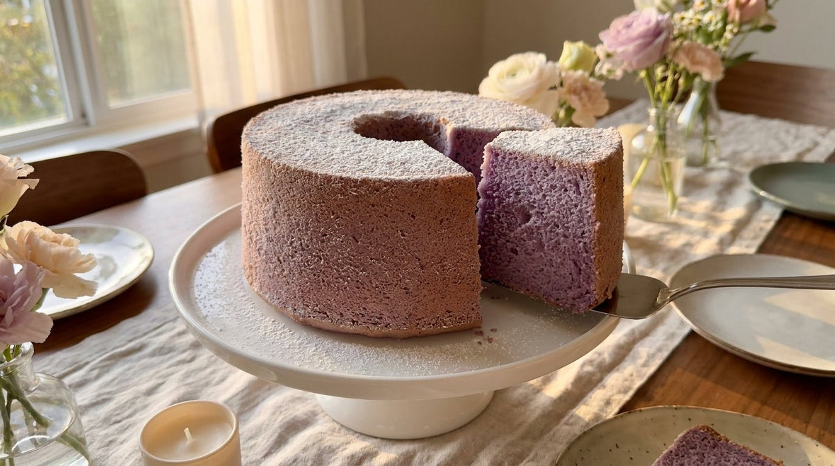 Tall ube chiffon cake on a white cake stand, naturally lavender with powdered sugar dusting and one slice cut showing the airy interior crumb