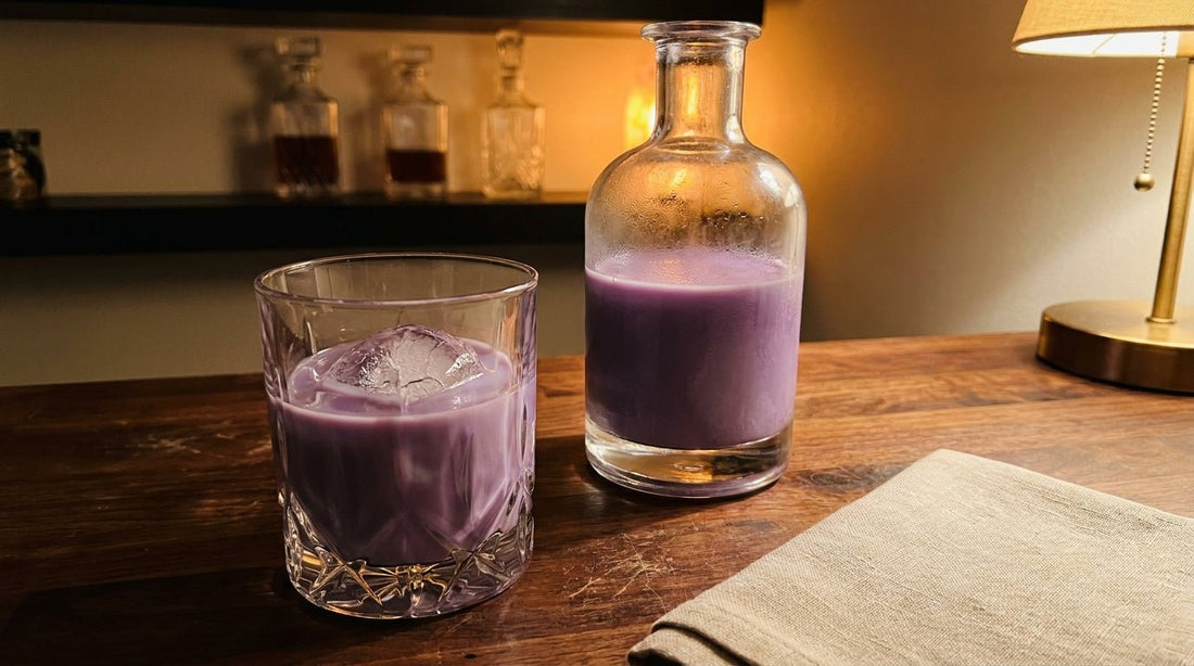 Glass of creamy lavender-purple ube cream liqueur over ice on a dark wooden home bar next to an unlabeled bottle