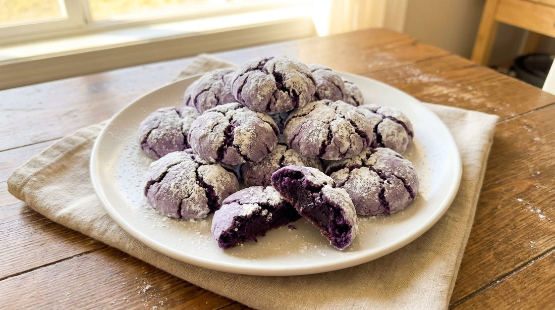 White plate piled with ube crinkle cookies — soft lavender cookies coated in white powdered sugar with dramatic dark purple cracks, one broken to show the fudgy interior