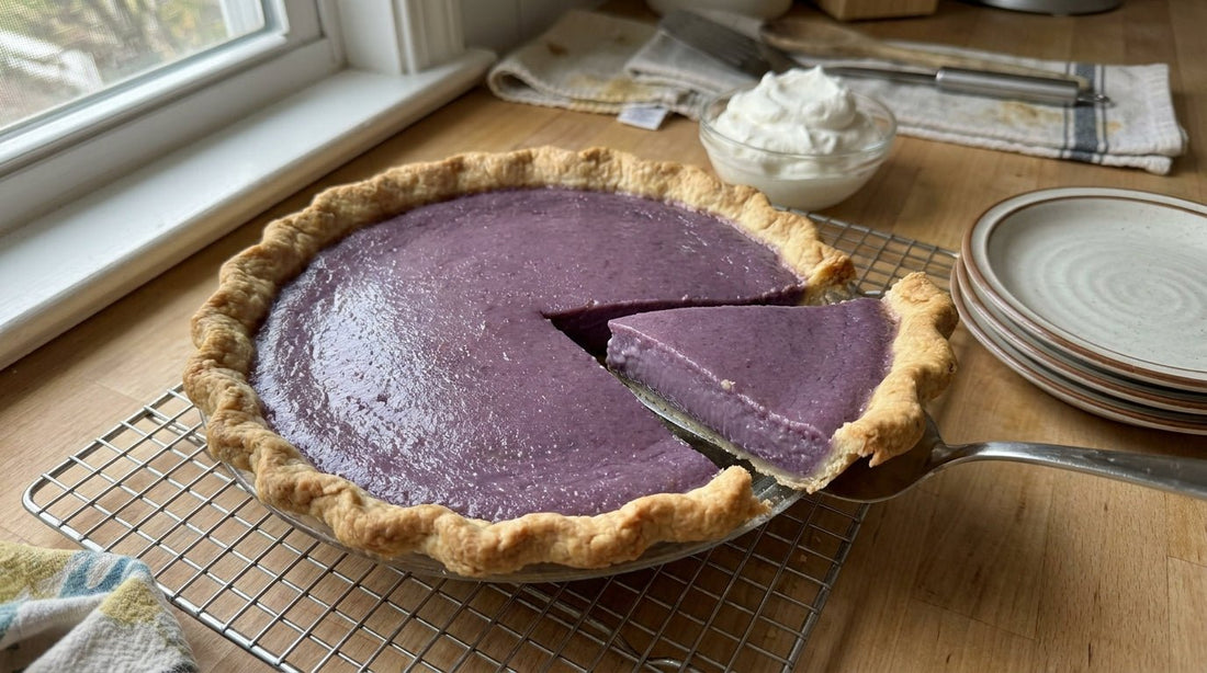 A whole 9-inch ube pie on a wire cooling rack on a home kitchen counter — golden-brown crimped crust, deep lavender-purple custard filling, with one slice partially lifted out