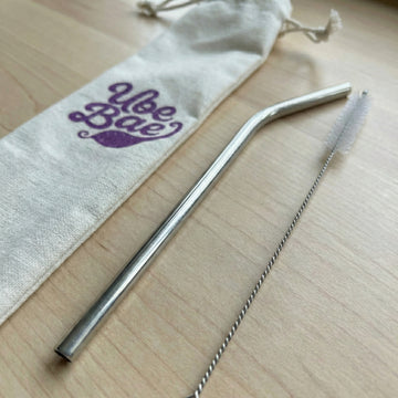 Metallic straw and cleaning brush on a wooden surface with a 'Ube Bae' branded drawstring bag.